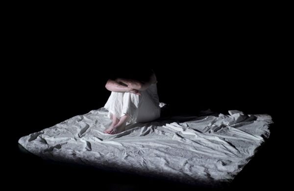A woman wearing white night clothes sits on a floor covered in a wrinkled white sheet. She ishugging her knees against her chest. She is half hidden in darkness with her face obscured.