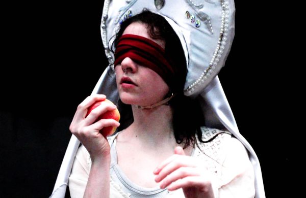 A woman dressed in white and wearing an elaborate hat. She is blindfolded with a scarf and is holding an apple close to her face.