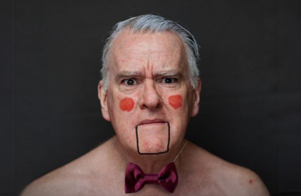 A photo of a grey haired man wearing make up and a bow tie that makes him look like a ventriloquist's dummy.