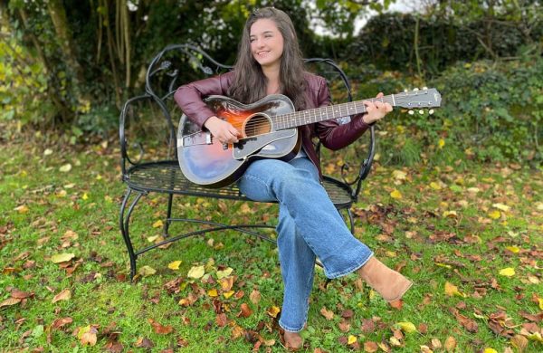 Image of a young white woman playing a guitar outdoors in a garden. She is sitting in a chair. Autumn leaves are strewn around her.